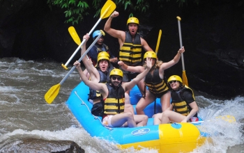 A group of tourists smiling and wearing life vests as they sit in a rubber boat on a river, with green trees and foliage in the background.